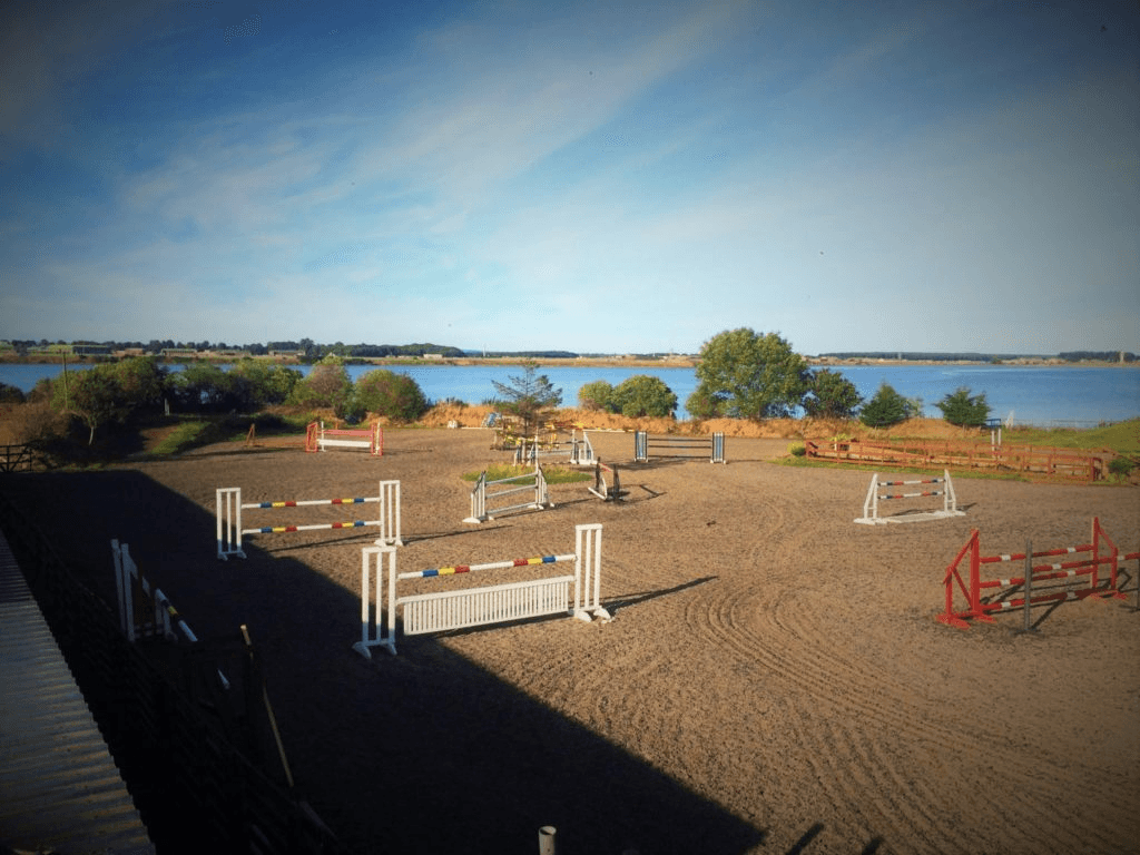 Colourful jumps in an outdoor arena with water and countryside beyond.